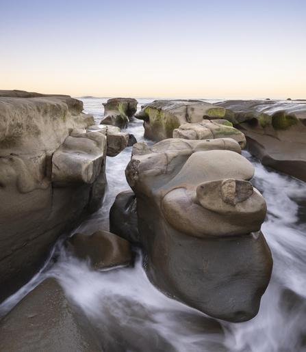 A color photograph of large rock formations along the coast of La Jolla, San Diego, California. Waves pass through the rocks as the sky lights up in warm tones at sunrise.