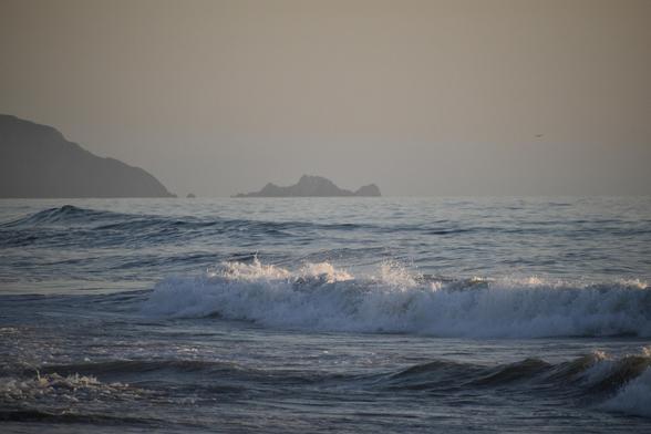 A beach scene with weird lighting. 

The sky is a hazy gray amber color, and some brown cliffs and outcropped rocks in the water at a distance are hazy and grayish. 

The surface of the water is a muted shiny light blue, with scattered dark shadows acting as ‘highlights’ since the sun is very low at the side of the image. 
One small crashing rolling wave up close at the shore is throwing up white splashes and drops that catch the low sunlight and are lit up at their tips and slightly pinkish.

Definitely a beach scene at sunset, but fairly surreal in its color and appearance.