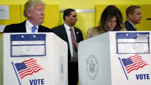 Donald Trump and Melania Trump stand at side-by-side voting booths in a polling place, with two men in suits behind them and yellow walls in the background. The booths display an American flag and the word VOTE.