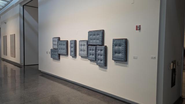artwork on the wall of a gallery... the artwork is a series of plush, upholstered cushions in a soft grey-blue color