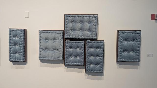 artwork on the wall of a gallery... the artwork is a series of plush, upholstered cushions in a soft grey-blue color and they are begging to be touched