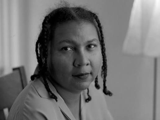 Black-and-white portrait of a woman with braided hair looking toward the camera, seated indoors. A lamp and part of a chair are visible in the background.