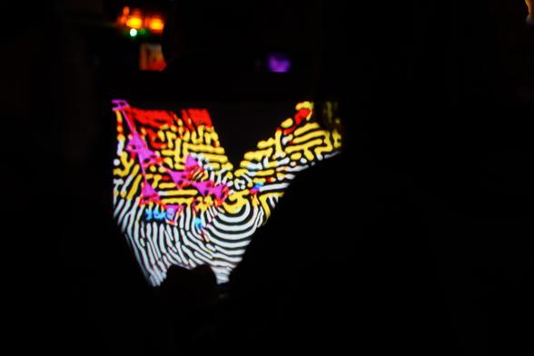 A player’s silhouette partly obscures a CRT running Hypnogenesis. Its screen shows curling dark and light bands, mostly white below and yellow above, accented with red, magenta and cyan. Small pools of orange, green and purple light break the blackness above the screen, their edges softened by blur.