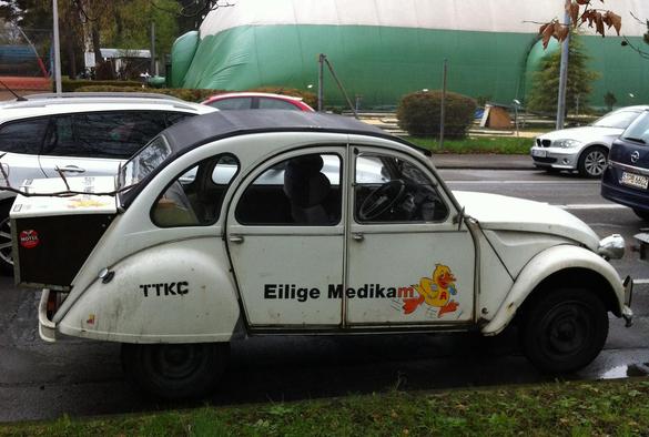 Ein weißer Citroën 2CV mit starken Gebrauchsspuren und einer Kofferraumvergrößerung. An der Seite über die Türen ein Schriftzug "Eilige Medikam-Ente" (wobei das Wort Ente durch die comichafte Darstellung einer Ente mit einem Apothekenlogo ersetzt ist).