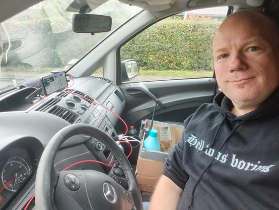 Me sitting in the cab of a Mercedes Vito. Walkie talking, GPS, snackbox and charging cables visible. And me in a hoodie with the text Hell was Boring.