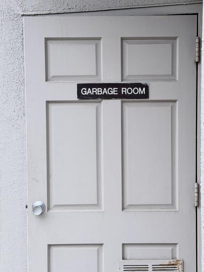 An off-white outside door with panels and a silver metal handle, with a white  stucco wall around it, slightly ajar. It has a reddish rusting slatted vent toward the bottom, and a small sign in the middle, near the top. 

It is a black sign with white block letters in a rounded typeface that reads ‘garbage room’.