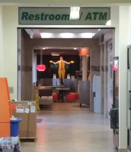 A strange, slightly unsettling photograph taken inside what appears to be a fast-food restaurant or highway rest stop.

At the top of the image, a large overhead sign reads “Restroom / ATM” in dark green letters.

Below the sign is a long, mostly empty hallway with beige walls, tiled floors, fluorescent ceiling lights, and various cardboard boxes, trash cans, and equipment along the sides.

At the very far end of the hallway, standing in a dimly lit dining area, is Ronald McDonald, the McDonald's clown mascot. He has bright red hair, a white-painted face, a yellow jumpsuit, red-and-white striped sleeves, and oversized yellow gloves.

His arms are stretched wide open, palms facing upward, as though he's dramatically welcoming someone, like buddy Jesus.

He's positioned directly in the center of the distant doorway, staring toward the camera. The darkness behind him makes his colorful costume stand out.

The humor: Someone apparently just wants to find the restroom or an ATM, but instead encounters Ronald McDonald standing ominously at the end of a deserted hallway with his arms spread wide.

The combination of the ordinary restroom sign, the eerie empty corridor, and the unexpectedly welcoming clown makes the scene feel like something out of a low-budget horror movie, or just another day in the USA.