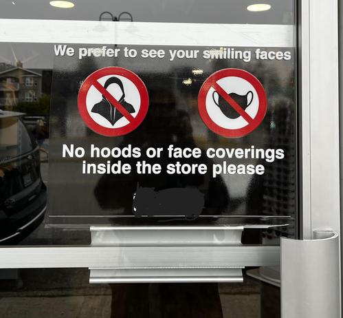 Store door that says “we prefer to see your smiling faces no hoods or face coverings inside the store please.” It has symbols for hoodies and medical masks with a big no symbol over them.