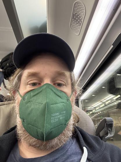 Selfie on a Eurostar train, 43 y/o white man with beard and long hair wearing a blue base cap and a green FFP2 mask
