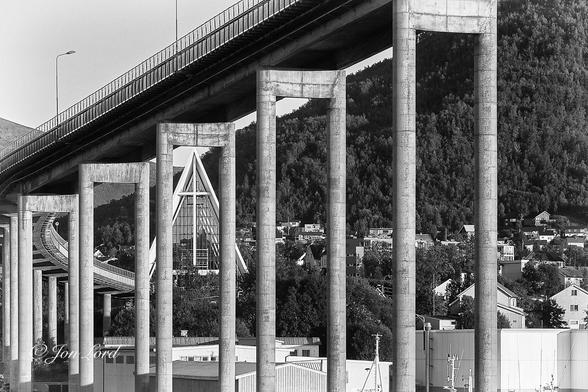 This is a black and white photo in landscape format, viewing through numerous pairs of tall concrete bridge supports with the Arctic Cathedral framed by one of the pairs. Tromsø, Norway (2017).
Stretching across the photo from right to left are the multiple (five in clear view with many more partially obscured) pairs of tall and thin cylindrical concrete columns supporting the bridge deck. The deck above is lined with a tall metal railing and with two tall street lights. Through the third pair of columns from the right is the Arctic Cathedral or correctly the Tromsdalen Church (Norwegian: Tromsdalen Kirke, Ishavskatedralen) often called the "The Cathedral of the Arctic Ocean". The camera is ten metres from the closest column and two or three hundred metres from the church. The church is viewed from the front with neither side in view. The white concrete front is shaped like a tall Isosceles triangle or ridge tent, with a base narrower than the two tall equal sides. In the centre is a thin crucifix that extends from the base to the apex with two cross members reaching outwards to the sides three-quarters of the way up. The remainder of the front is filled with horizontal rectangular frames of glass. In the foreground are the roofs of various small industrial buildings with the masts of one fishing vessel visible in the lower right corner. Stretching across the centre level and behind the church are numerous residential houses with a dark, tree covered mountain close behind and sloping steeply downwards from the upper right corner towards the lower left. Above is a sky overexposed to a pale white, devoid of any detail.