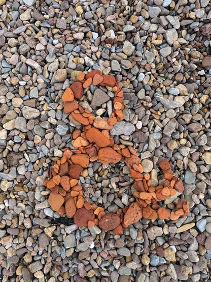 An ampersand formed by arranging reddish stones on a pebbled beach.