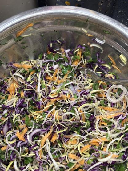 A metal bowl filled with finely sliced purple cabbage, leeks, carrots, and some herbs all tossed together. There is no dressing yet.