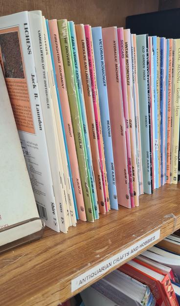 A bookshelf in a charity shop labelled "Antiquarian crafts and hobbies". It's filled with a few dozen very thin books with titles including "Lichen", "Umbellifers of the British Isles",  "Sidecars", "Spinning and Spinning Wheels" and "Old Garden Tools".