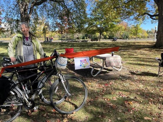 Photograph of a perk setting with two bicycles and a smiling white woman holding her bike with a small petty flag at the midpoint of an Orange pole with a picnic table in the background.