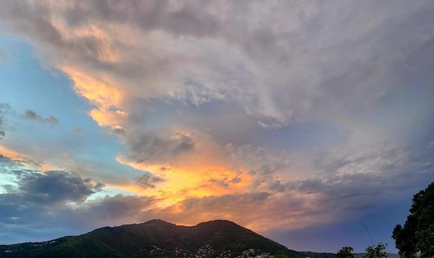 A dramatic sunset over a dark, green mountain ridge, with brilliant orange and golden light illuminating layers of swirling gray and purple clouds. Patches of turquoise-blue sky contrast with the stormy cloud cover, while scattered houses dot the foothills and trees frame the right edge.