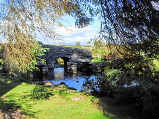 The East Dart River at Bellever, Dartmoor.
A stone bridge viewed through overhanging branches.  The bridge is supported by stone arches, one of which is clearly visible.
In front of the bridge is one span of an incomplete clapper bridge: a granite slab supported by the bank at one end and a granite pile at the other.
The bridges and the blue sky are reflected in the river, and an area of short green grass takes up the foreground.