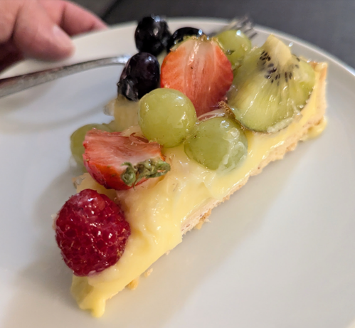 A photo shows what looks like a slice of pie, but it's a tart pastry filled with yellow custard. On top are fresh fruits: Raspberries, strawberries, grapes, kiwi slices and blueberries.