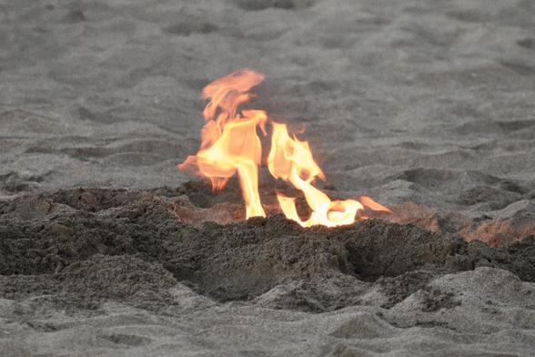 In the middle of some light gray beach sand, a small ditch dug with darker gray sand around it hosts a little fire burning. The wood fuel isn’t visible, just the golden and orange flames licking up out of the pit in twisting and jagged patterns. The insides of the fire pit are also glowing a little red from the firelight reflected on the dark gray sands.