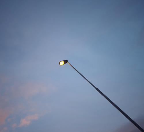 A black street lamp, photographed from below, stretching diagonally from the bottom right-hand corner towards the centre of the picture and already lit, set against a pastel-blue sky with a few pink clouds in the bottom left-hand corner.
