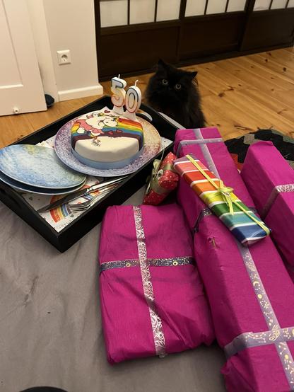 A colorful birthday setup featuring a decorated cake with a "50" candle, surrounded by several wrapped gifts in pink and other colors. A black cat is nearby, and the scene is set on a light-colored surface with additional plates and utensils visible.