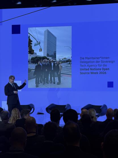 Erik Möller steht links auf einer Bühne und gestikuliert während einer Präsentation. Im Hintergrund ist eine große blaue Leinwand zu sehen, auf der ein hochkant arrangiertes Foto gezeigt wird. Das Foto stellt eine Gruppe von Menschen vor dem Hauptgebäude der Vereinten Nationen in New York an einer Straßenecke dar. Rechts neben dem eingeblendeten Foto steht folgender deutscher Text auf der Leinwand: „Die Maintainer*innen-Delegation der Sovereign Tech Agency für die United Nations Open Source Week 2026“. Im Vordergrund schaut das Publikum von hinten auf die Bühne.