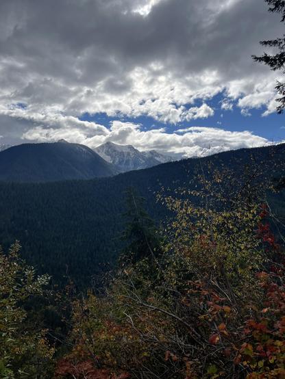 A cloudy sky, blue and white, over a snowy peak. Next, forested mountains, then red and yellow deciduous trees.