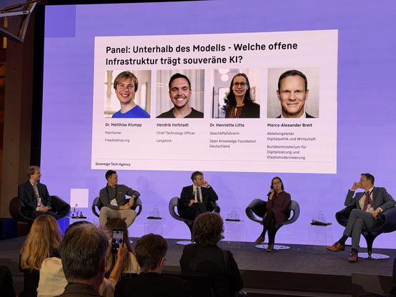 Foto von einer Podiumsdiskussion „Panel: Unterhalb des Modells – Welche offene Infrastruktur trägt souveräne KI?“. Auf einer großen Leinwand im Hintergrund sind die Porträts und Namen von vier Teilnehmenden zu sehen: Dr. Matthias Klumpp (Maintainer, Freedesktop.org), Hendrik Hofstadt (Chief Technology Officer, Langdock), Dr. Henriette Litta (Geschäftsführerin, Open Knowledge Foundation Deutschland) und Marco-Alexander Breit (Abteilungsleiter Digitalpolitik und Wirtschaft, Bundesministerium für Digitalisierung und Staatsmodernisierung). Auf dem Podium davor sitzen vier der Personen in modernen Sesseln auf einer Bühne und diskutieren, während eine Person mit Mikrofon spricht. Im Vordergrund sind verschwommen die Köpfe und Schultern des Publikums zu sehen. Das Logo der „Sovereign Tech Agency“ ist unten links auf der Leinwand zu erkennen.