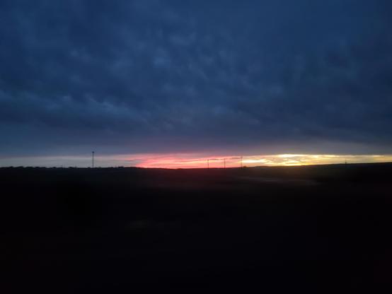 A photo of a sunrise. The bottom half of the photo is almost entirely black flat landscape. The top half of the photo is a dark blue sheet of clouds. A sliver at the horizon is open sky. On the left third is cloudy and grey. The center third is a grapefruit orange red color, while the right third is a beautiful golden.