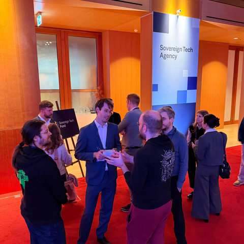 Eine Gruppe von Menschen steht in einem Foyer auf rotem Teppich und unterhält sich. Im Hintergrund ist ein Aufsteller der „Sovereign Tech Agency“ zu sehen.