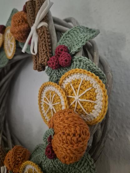 A close-up of a charming hand-crocheted wreath bursting with autumn warmth. Crocheted orange slices in bright shades of yellow and orange sit alongside brown cinnamon sticks tied neatly with a white ribbon, while green leaves and plump red berries fill the gaps between them. The decorations are attached to a light-colored grapevine base, giving the piece a natural, rustic texture. Every element is lovingly handmade, with the vibrant yarn colors evoking the cozy scents and hues of fall and winter — oranges, spice, and berries gathered in one decorative circle ready to welcome the season.
