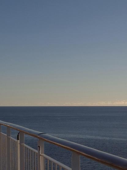 Over a railing that runs diagonally from the bottom right-hand corner to the left, looking out over a tranquil blue sea that meets a light blue sky on the horizon.