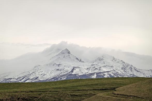 Paretaitonga summit---part of the Ruapehu complex. The summit is white, the cloud behind it is darker white, and the sky is brighter white. Olive green grass below.
