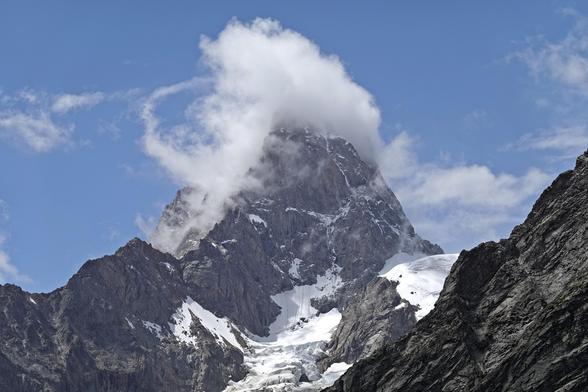 A narrow pointy rocky mountain in the Alps with a glacier descending from its flanks. Again there is a cloud wrapped around the top of the mountain.
