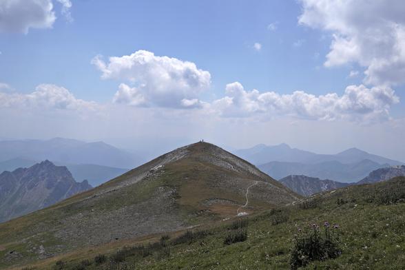 A small roundish peak with two people descending Korab and heading to Albania. Other mountain ranges visible in the distance.