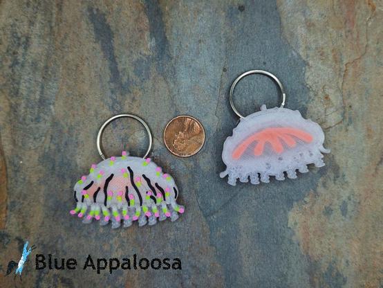 Two 3d printed Flower-hat Jelly designs. On the left, a jelly-fish like creature with translucent bell and coiled tentacles. The bell has vertical black stripes and short tentacles tipped with neon green and pink. The left design shows the “back” of the design, with an orange inner structure visible through the translucent body. Both designs have-inch (2.5cm) split ring through the top of the bell. In the middle is a US penny for scale.
Watermark reads: Blue Appaloosa