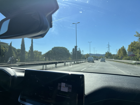 Udsigt gennem en bils forrude. Man kan se motorvej mens også cypresser, andre træer og blå himmel