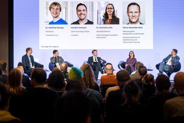 A panel discussion on a stage featuring five participants sitting in armchairs. The large screen behind them displays their portraits, names, and titles, while the audience listens in the foreground.