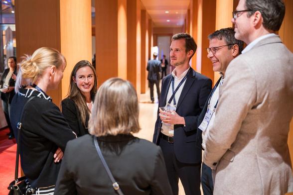 A group of five people standing together in a warmly lit corridor, chatting and smiling during an event break. They are wearing "Sovereign Tech Agency" name tags.