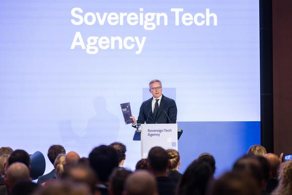 Karsten Wildberger in a dark suit standing at a "Sovereign Tech Agency" lectern on a stage in front of an audience. He is holding up a brochure and speaking into microphones. A large screen in the background displays the text "Sovereign Tech Agency".