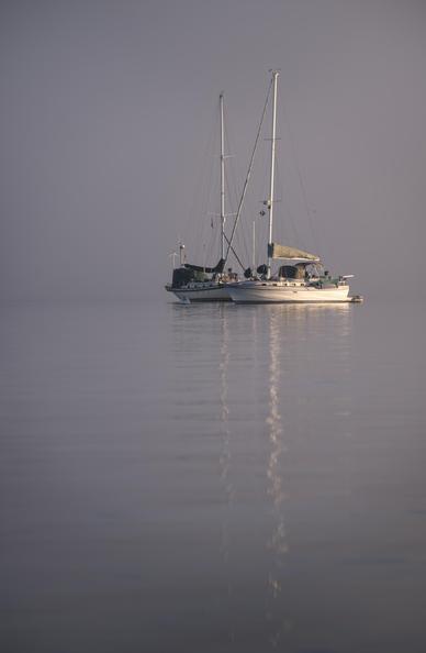 A color photograph of morning fog burning off of San Diego Bay, California. Two boats can be seen anchored in the water with a thick wall of clouds behind them.
