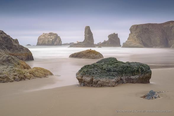 Color seascape photo showing large rocks at a beach and in the surf. The water is rendered smooth with a slow shutter speed. A gentle mist hangs in the air and the atmosphere is peaceful, calm, and perhaps a bit mysterious. Two birds can be seen on a rock in the surf.