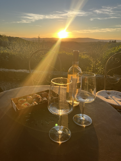 Terrassebord og -stole i smedejern foran en bedårende solnedgangsudsigt over toscanske bakkedrag. På bordet står en æske profiteroles, en flaske hvidvin og to glas. Stråler af modlys øger blot romantikken