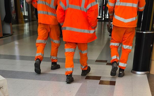 Three men walking through an indoor shopping centre. They are all wearing full orange hi-vis all-weather clothing, commonly seen on construction workers and the like in the UK. The picture cuts off at their upper backs so they're in no way identifiable, and the logos of their construction companies aren't visible either.