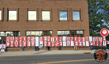 Outside the Trump rally, protestors are holding up a banner that says "BARRON'S MENTOR ANDREW TATE CHARGED W HORSE AND EEL PORN!"