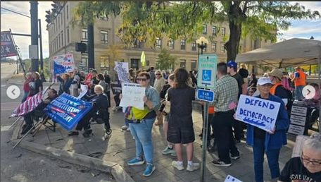 More of the crowd of protestors can be seen standing on the sidewalk with signs like "DEFEND DEMOCRACY" with a blue wave at the bottom. There is a tent set up and safety marshalls are seen in hi vis vests.