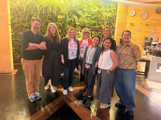 Eight members of the AoIR Executive Committee pose together indoors. They are wearing conference lanyards and badges, with warm lighting, wood-paneled walls, and a café counter in the background.