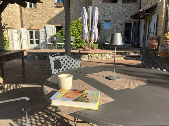 Terrassebord med stole omkring. I baggrunden et hus bygget af grove, gule sten i to etager med en hævet stenterrasse foran. På bordet står en lampe og en kop kaffe. Bogen Yesteryear af Caro Claire Burke pynter også