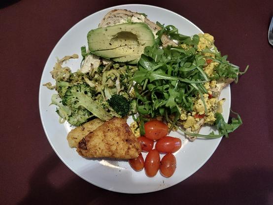 A white plate containing toast, one piece topped with smashed avocado and the other,  scrambled tofu, stir fried greens (broccoli and cabbage), fried mini plum tomatoes and hash browns, tower with plenty of rocket. All cooked by my step they.