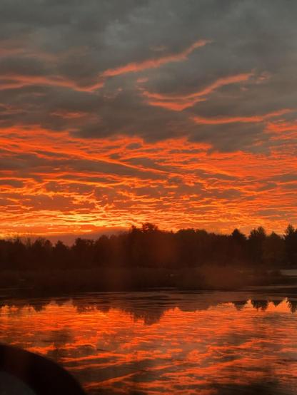 The sky is a fiery mix of pink and orange glowing from the bottom of the clouds, reflected in the still lake and separated by a black silhouette of the tree line.