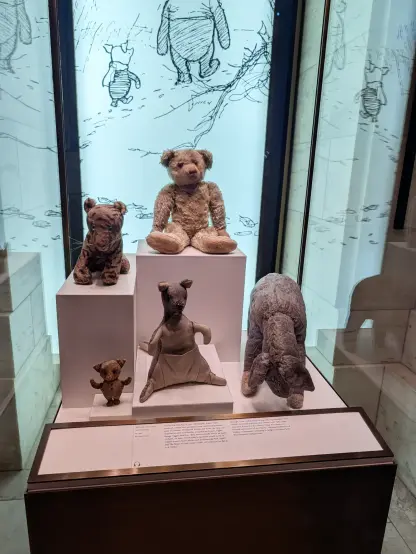 Photo of a museum display of stuffed animals encased in glass. A teddy bear sits on the tallest box with a tiger on the next-tallest (to our left). The bottom row has a tiny pig, a kangaroo, and a donkey.

The placard reads:

Winnie-the-Pooh and Friends
ca. 1921
Rare Book Division

On his first birthday, in 1921, Christopher Robin Milne received a teddy bear purchased from Harrods department store in London. Christened Winnie-the-Pooh, the bear soon acquired several now-familiar companions: Eeyore, Piglet, Kanga, Tigger, and Roo. (Roo was eventually lost in an apple orchard.) In time, Christopher's playmates would come to inspire several classic works such as "Winnie-the-Pooh" (1926) and "The House at Pooh Corner" (1928), all written by his father, A.A. Milne.

Brought to the United States in 1947, the toys remained with Milne's American Publisher, E.P. Dutton, until 1987, when they were donated to the Library. Today they stand as a beloved centerpiece of the Library's renowned collection of children's literature, continuing to delight and inspire both the young and young at heart.